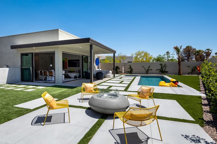 The backyard in full sun: yellow chairs around the fire pit, the covered patio and the lap pool