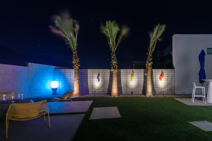 The backyard at night, with palms uplit and colored lights along the wall