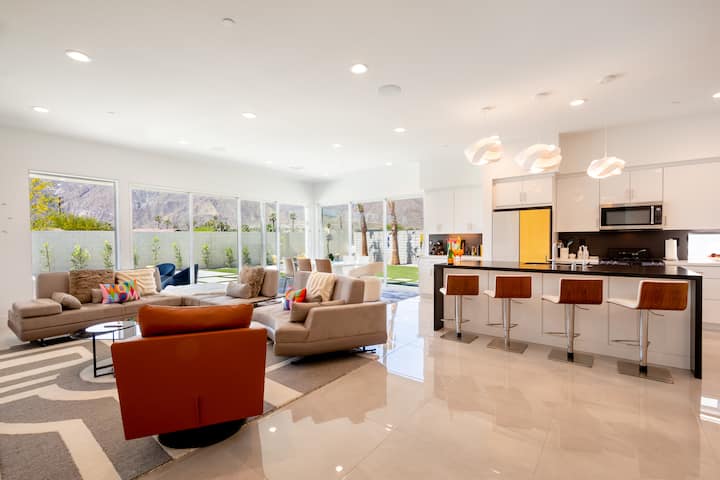 The great room, with sofas and an orange swivel chair, open to the kitchen and its island with four stools