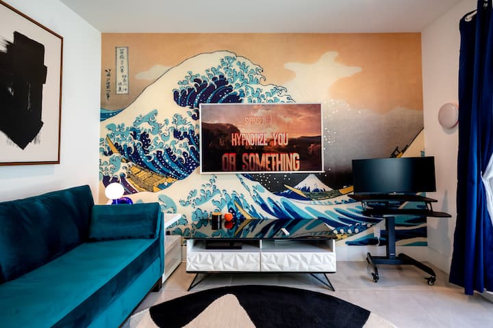 Guest room with a teal sofa, a desk with a monitor and a wall-size wave mural