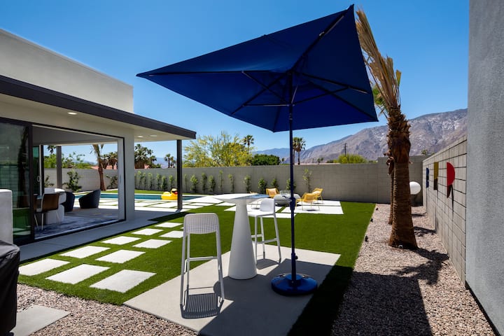 A high-top table and chairs under a blue umbrella beside the house